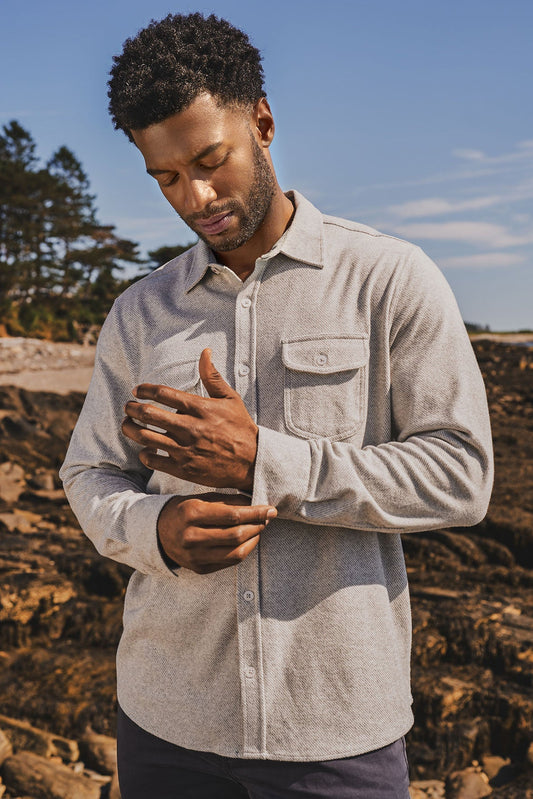 A man wearing a light gray long-sleeve twill flannel button-up shirt with a chest pocket, standing outdoors on a rocky shoreline under a blue sky.