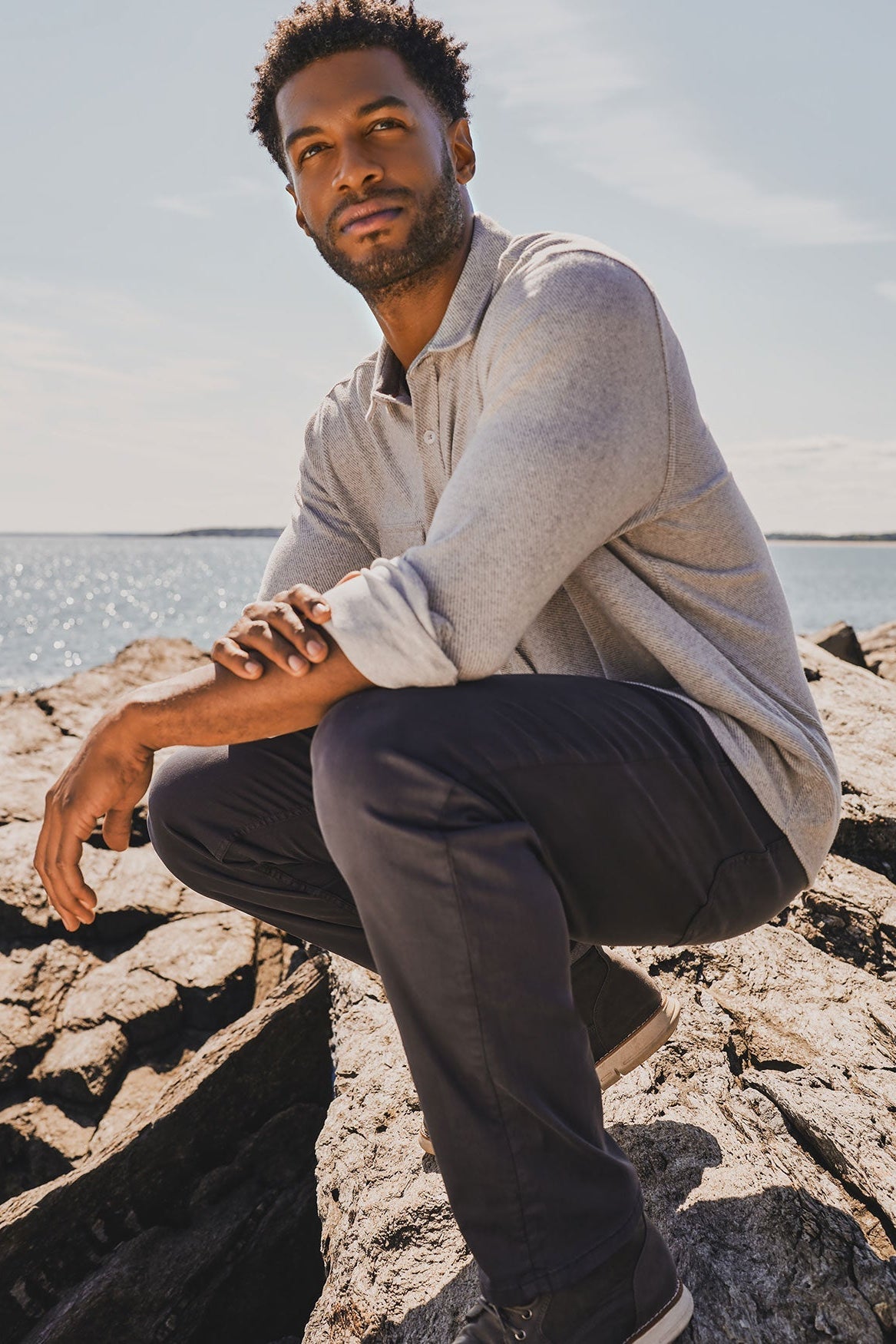Man sitting on rocks by a body of water wearing a light gray twill flannel and dark pants.