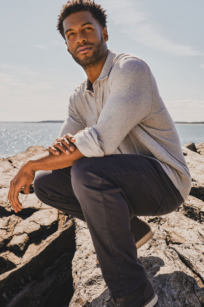 Man sitting on rocks by a body of water wearing a light gray twill flannel and dark pants.
