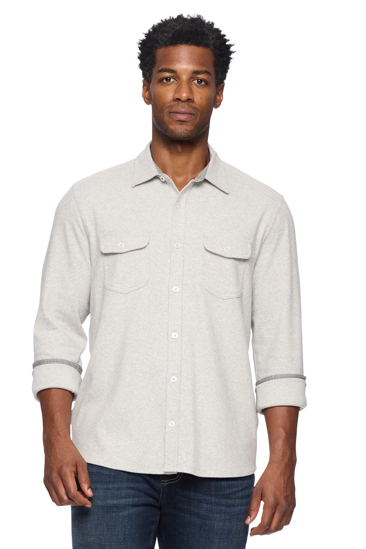 A man wearing a light heather gray twill long-sleeve button-up flannel shirt with two chest pockets and rolled cuffs, paired with dark blue jeans, against a plain white background.