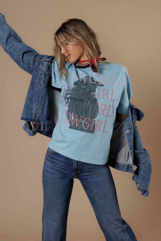 A woman modeling a light blue graphic t-shirt with a black cowgirl print and pink text, styled with a denim jacket draped off her shoulders and blue jeans, against a solid beige background.
