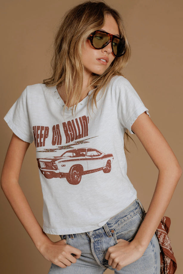 A woman wearing an ice blue graphic t-shirt with a brown distressed print of a muscle car and the text 'KEEP ON ROLLING', paired with light-wash jeans and sunglasses, against a solid tan background.