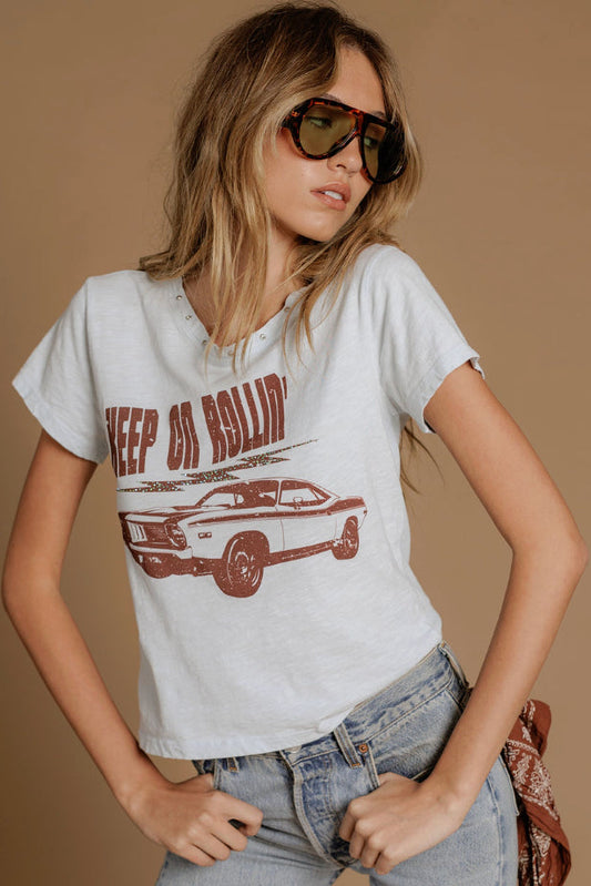 A woman wearing an ice blue graphic t-shirt with a brown distressed print of a muscle car and the text 'KEEP ON ROLLING', paired with light-wash jeans and sunglasses, against a solid tan background.