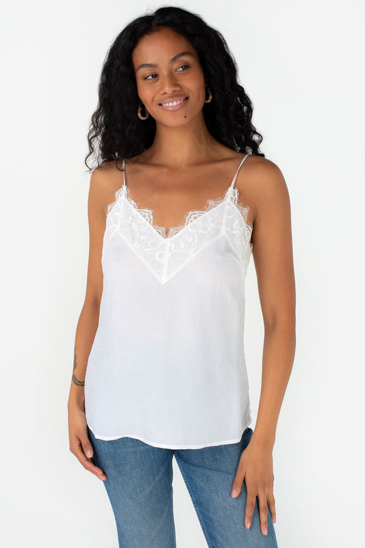 A woman modeling a chalk white spaghetti-strap camisole top with a delicate lace V-neckline, paired with blue jeans, against a plain white background.