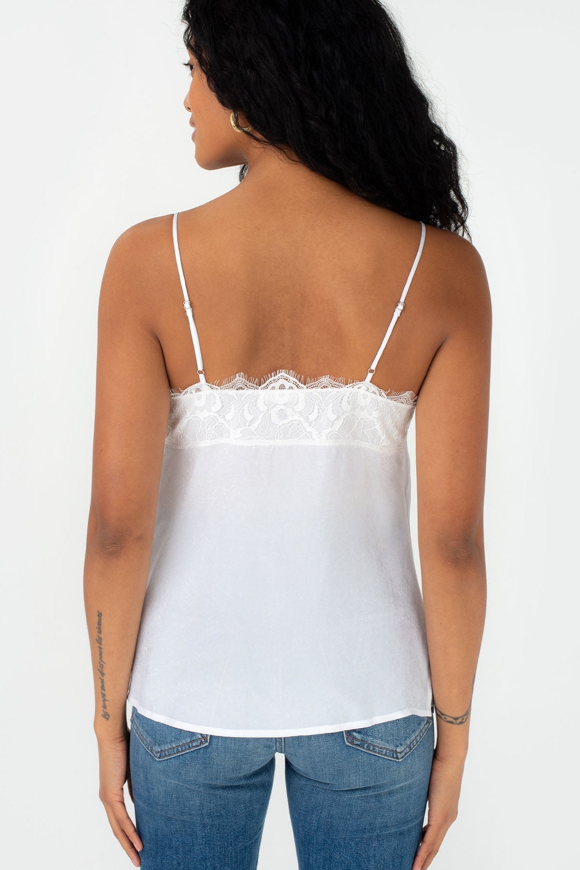 A woman shown from the back wearing a chalk white spaghetti-strap camisole top with a lace band across the upper back, paired with blue jeans, against a plain white studio background.