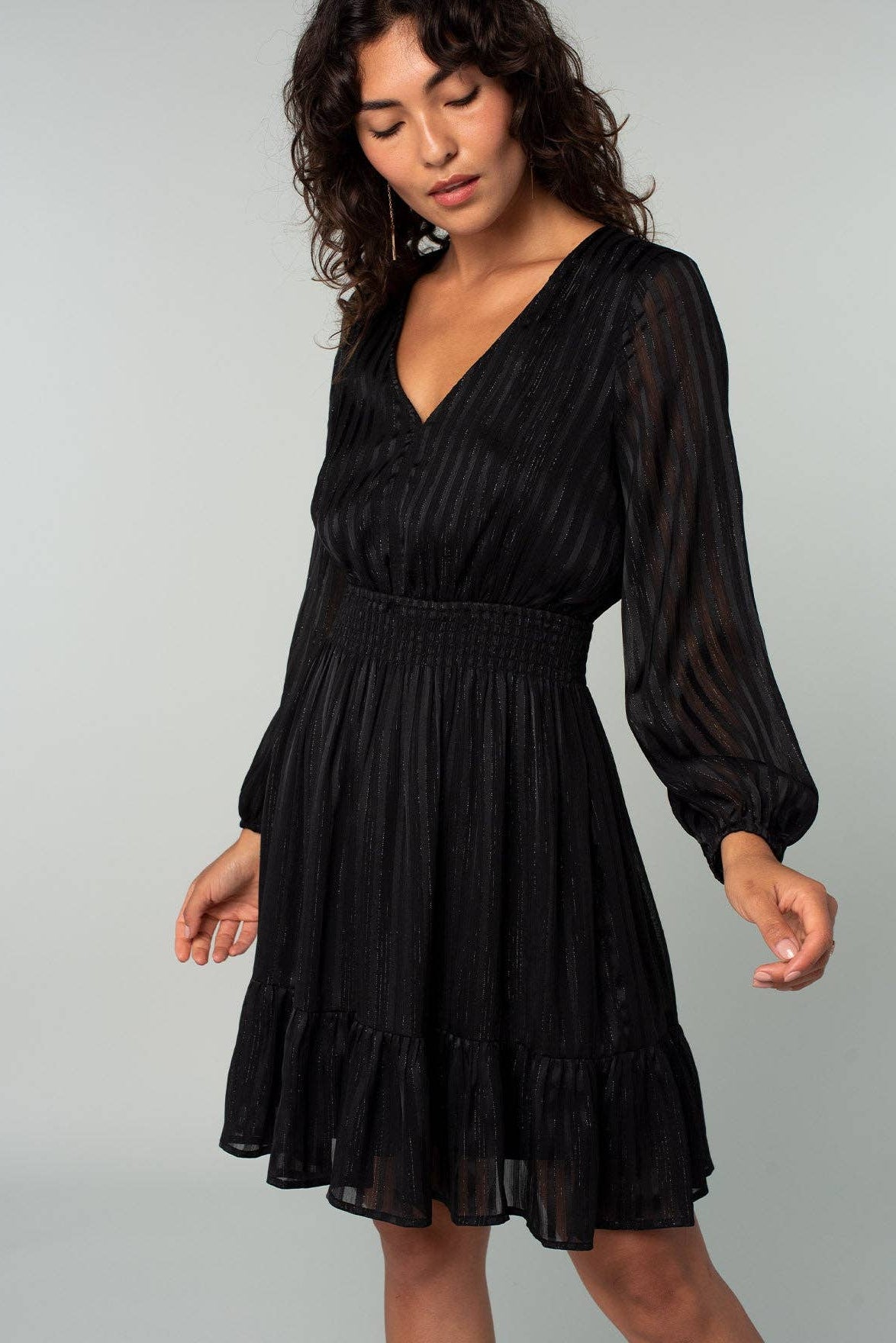 A woman wearing a black long-sleeve mini dress with a smocked waist and tiered skirt, photographed against a plain light gray studio background.