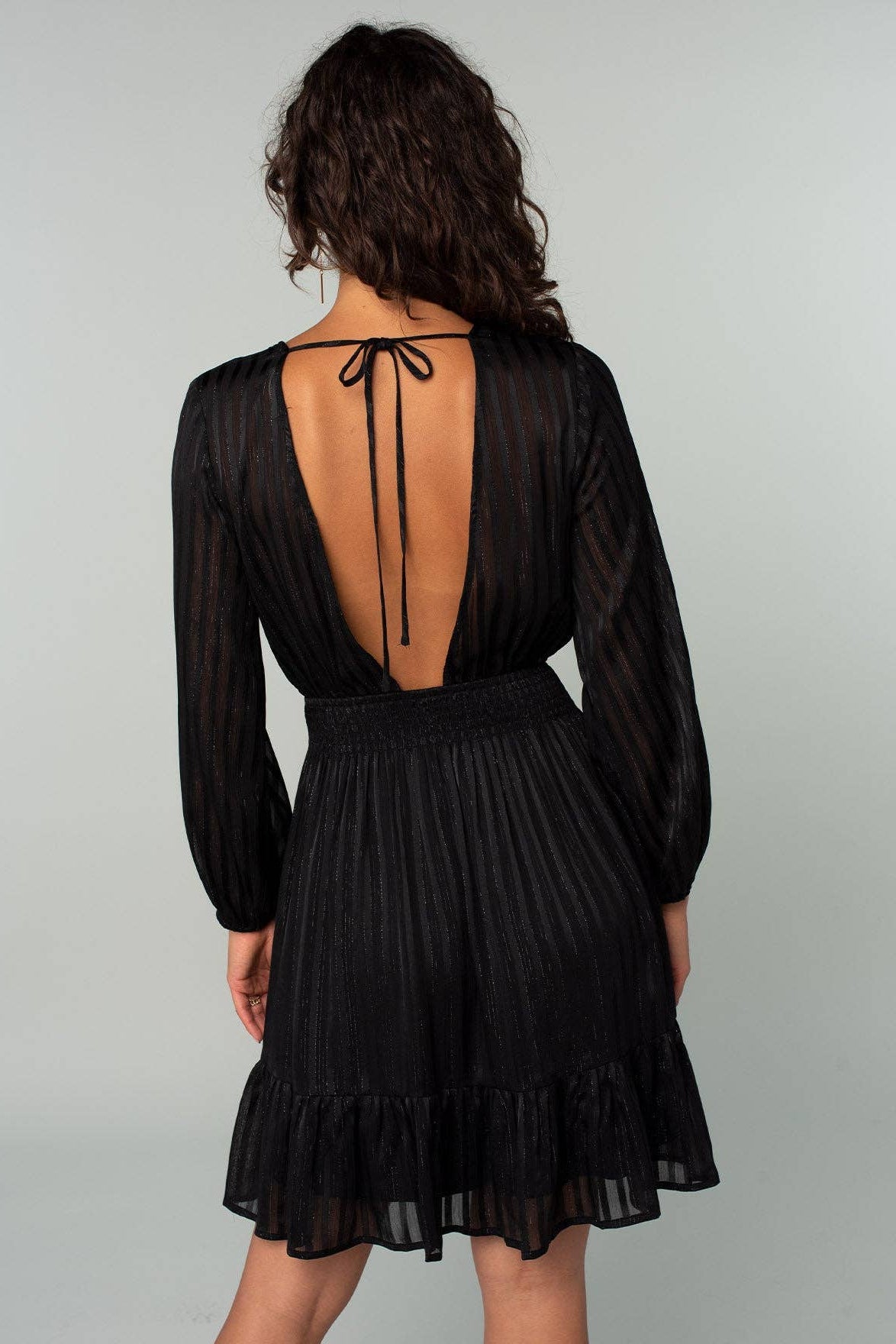 A woman shown from the back wearing a black long-sleeve midi dress with a sheer open back, tie detail, and ruffled hem against a light gray studio background.