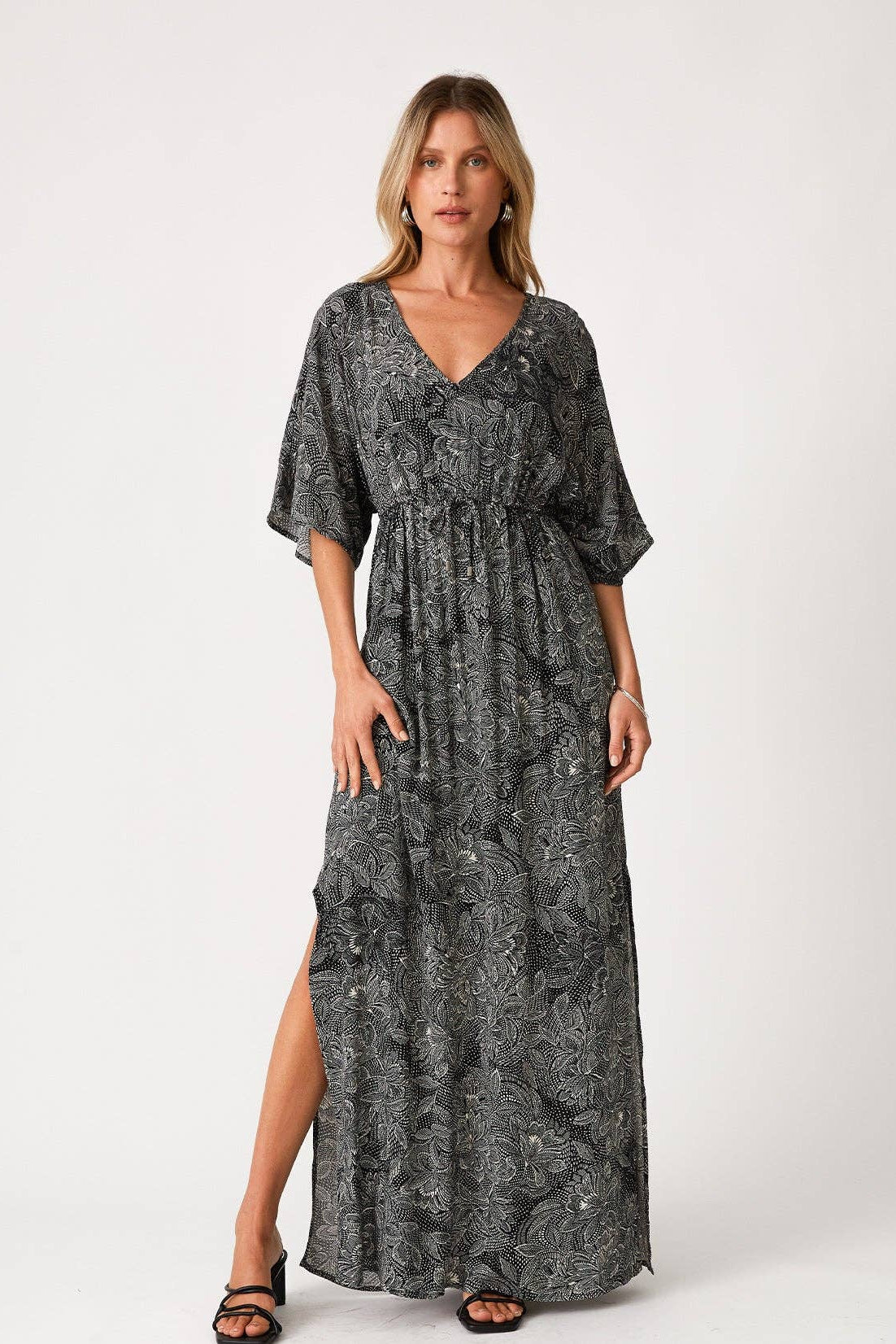 A woman modeling a dark gray and black floral print maxi dress with a V-neckline, short flutter sleeves, and a side slit, photographed against a plain white studio background.