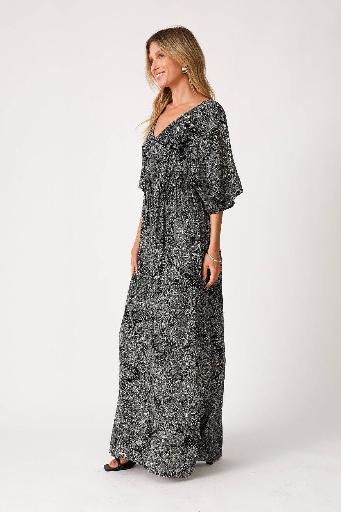 A woman modeling a long, black & natural maxi dress with a light gray abstract floral pattern, featuring a V-neckline, ruffled sleeves, and a tie at the waist, set against a plain light gray studio background.