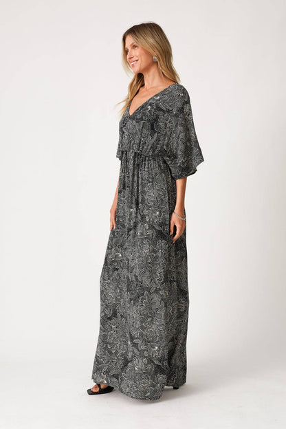 A woman modeling a long, black & natural maxi dress with a light gray abstract floral pattern, featuring a V-neckline, ruffled sleeves, and a tie at the waist, set against a plain light gray studio background.