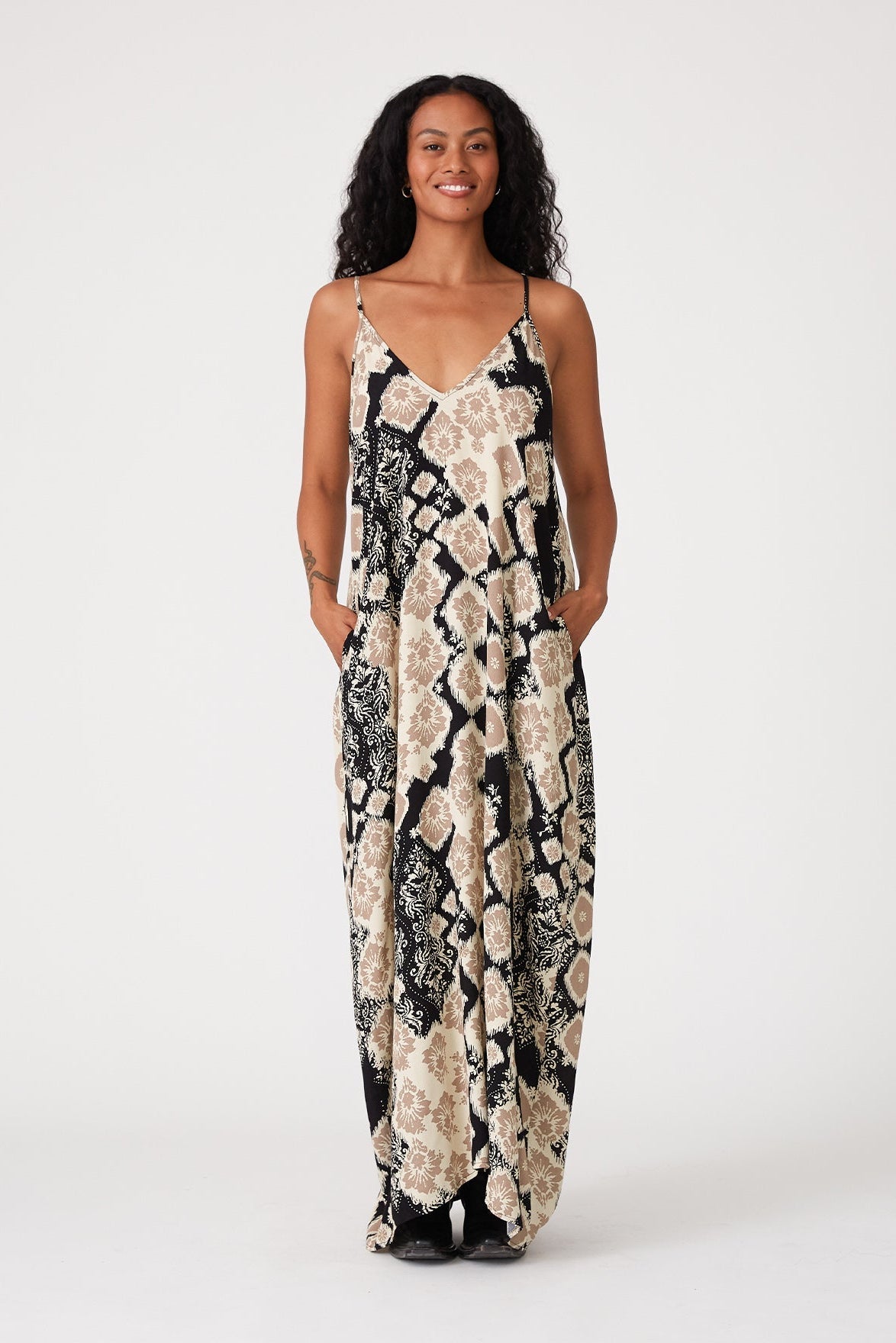 A woman modeling a sleeveless maxi slip dress with thin straps and a black, khaki, beige, and white abstract pattern, standing against a plain white studio background.