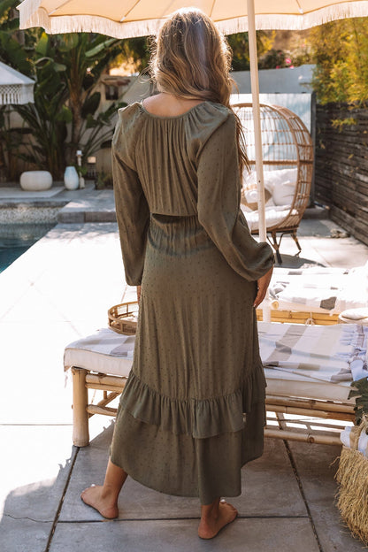 A woman stands barefoot by a poolside patio wearing a long-sleeved olive green maxi dress with a tiered ruffle hem, photographed from behind.