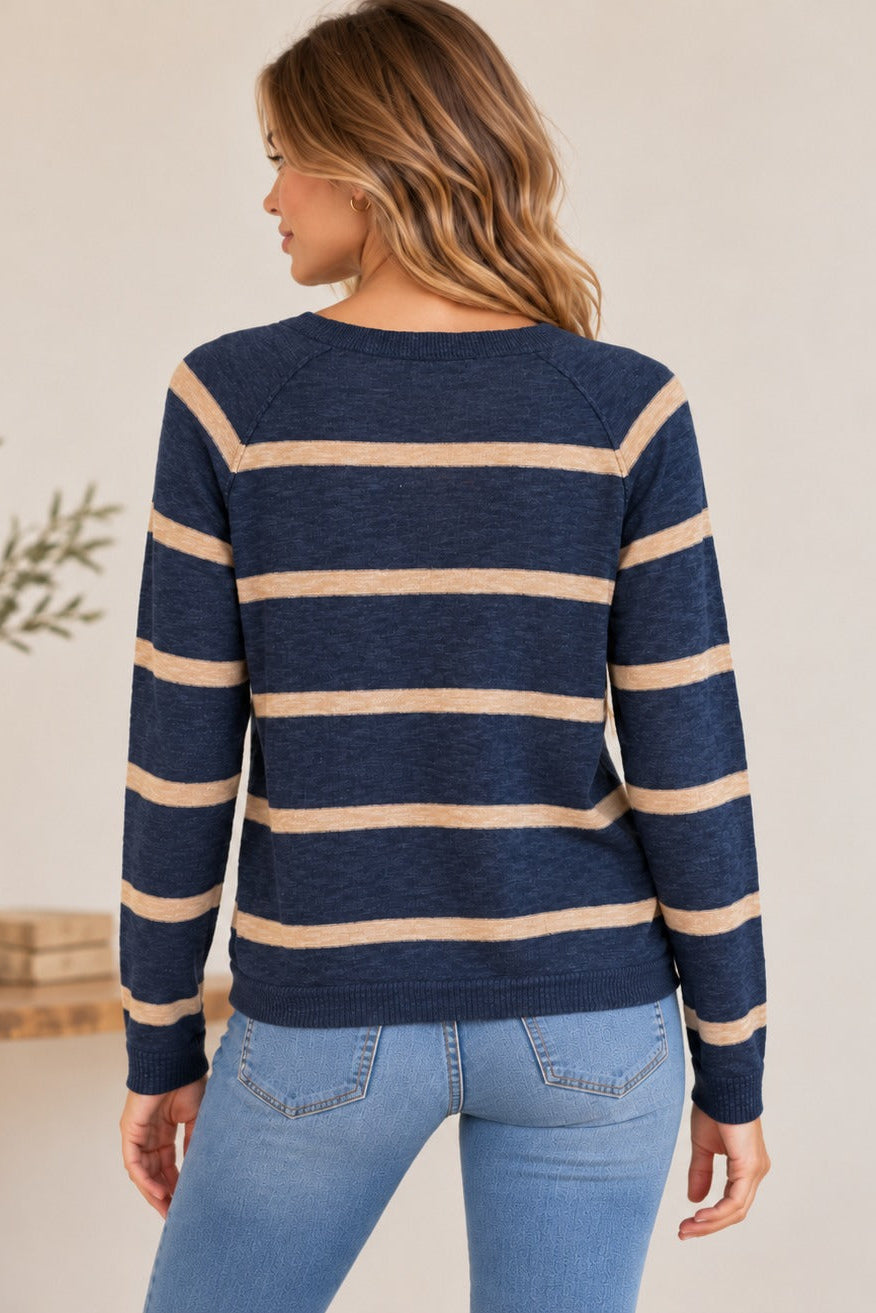 A woman shown from the back wearing a midnight blue and beige striped knit sweater with light blue jeans, standing in a softly lit indoor setting.