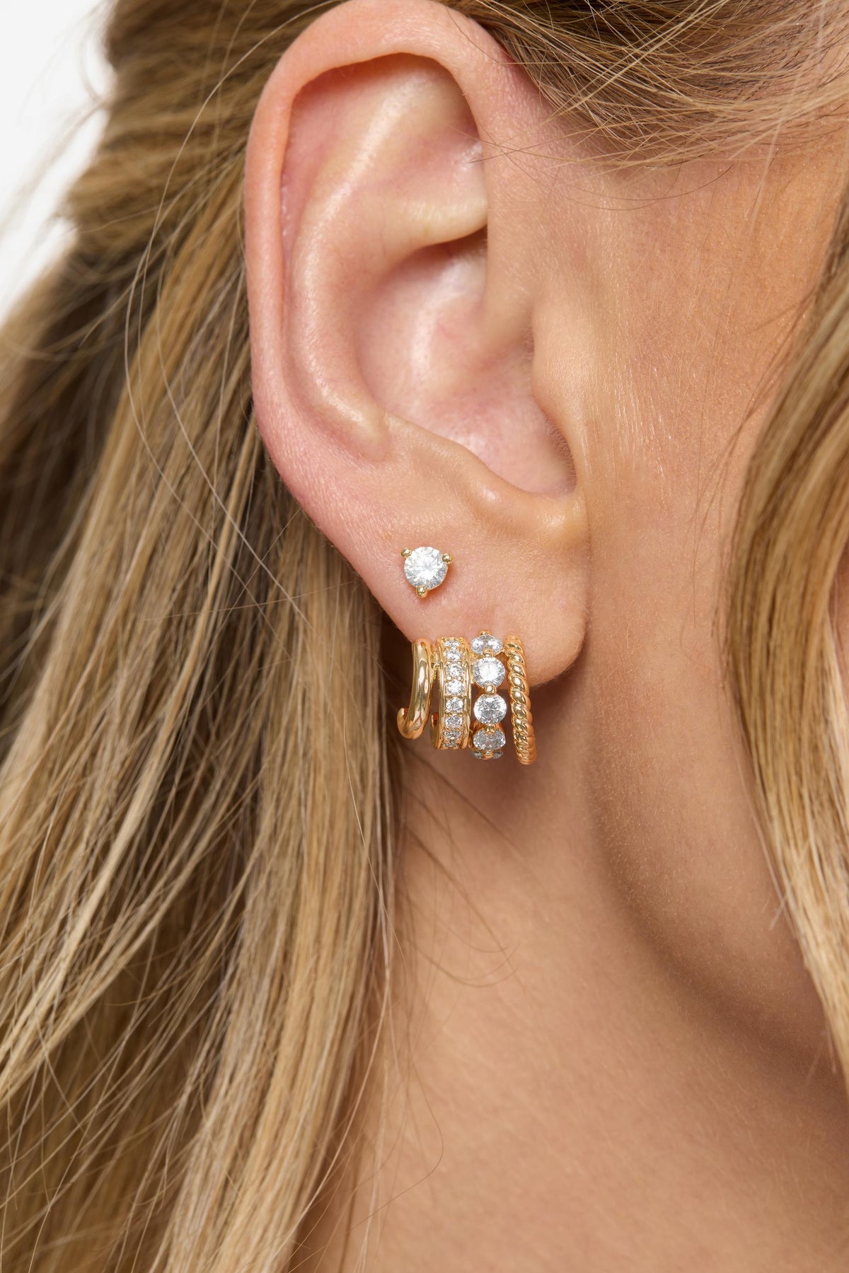 A close-up of a model's ear wearing a pair of gold huggie hoop Ashley Venice earrings adorned with clear crystals and a small matching crystal stud earring.
