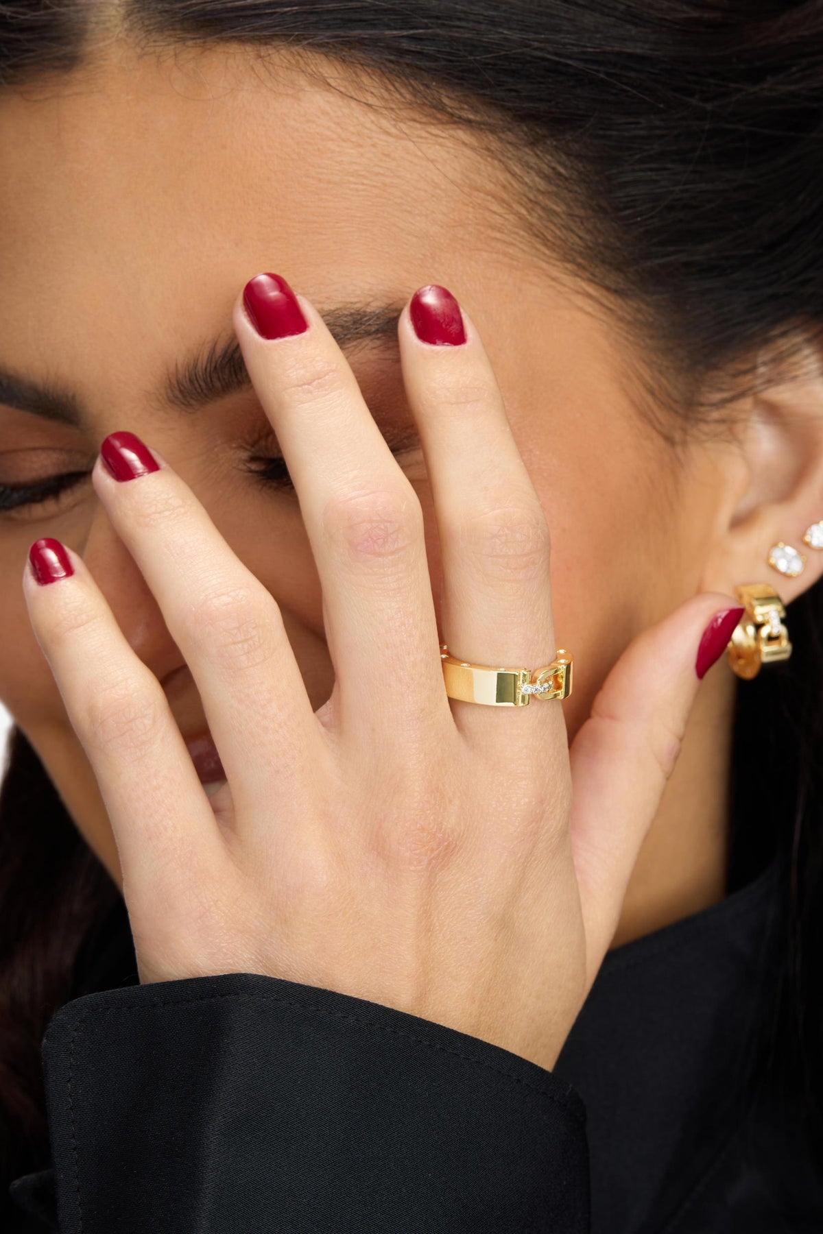 A close-up of a woman's hand with dark red manicured nails wearing an Ashley Smooth Lock wide gold band ring, with matching gold and crystal stud earrings visible on her ear. She is wearing a black top against a light gray background.