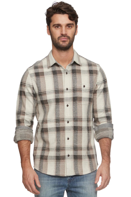 A man wearing a long-sleeve plaid button-up shirt in cream and charcoal tones, paired with blue jeans, against a plain white background.