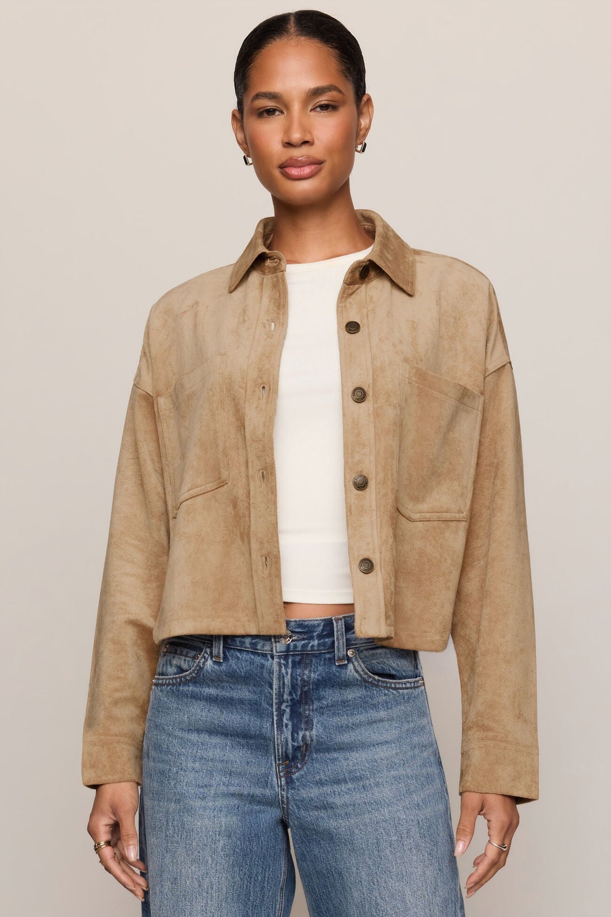 A woman modeling a cropped light sand suede jacket over a white ribbed top and blue jeans, photographed against a plain light beige studio background.