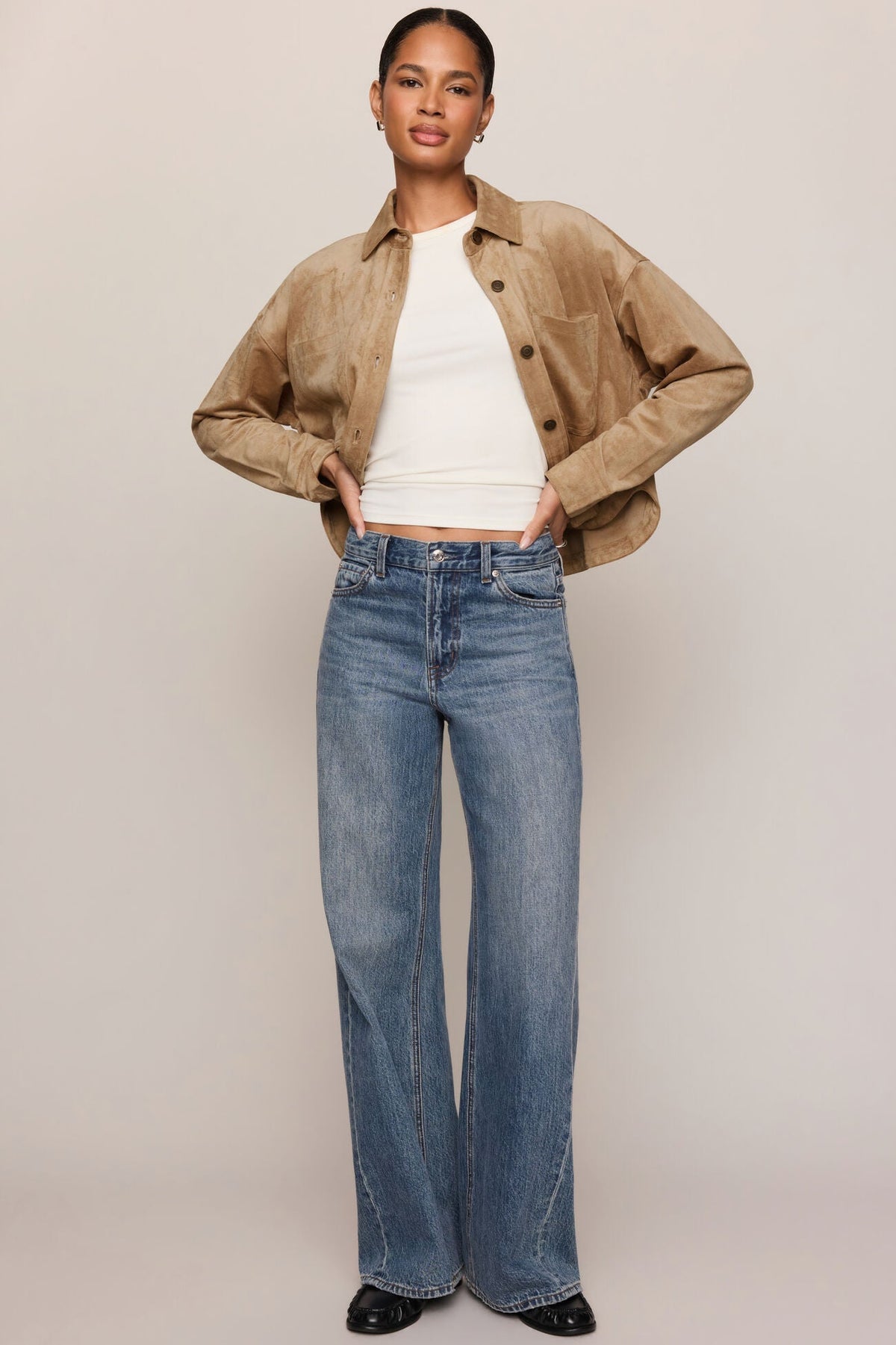 A model stands against a light beige studio background wearing a cropped light sand suede jacket over a white ribbed top and high-waisted, wide-leg blue jeans.