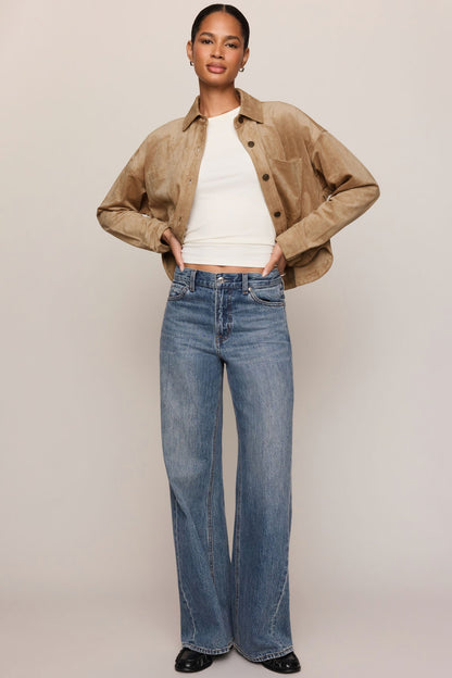 A model stands against a light beige studio background wearing a cropped light sand suede jacket over a white ribbed top and high-waisted, wide-leg blue jeans.