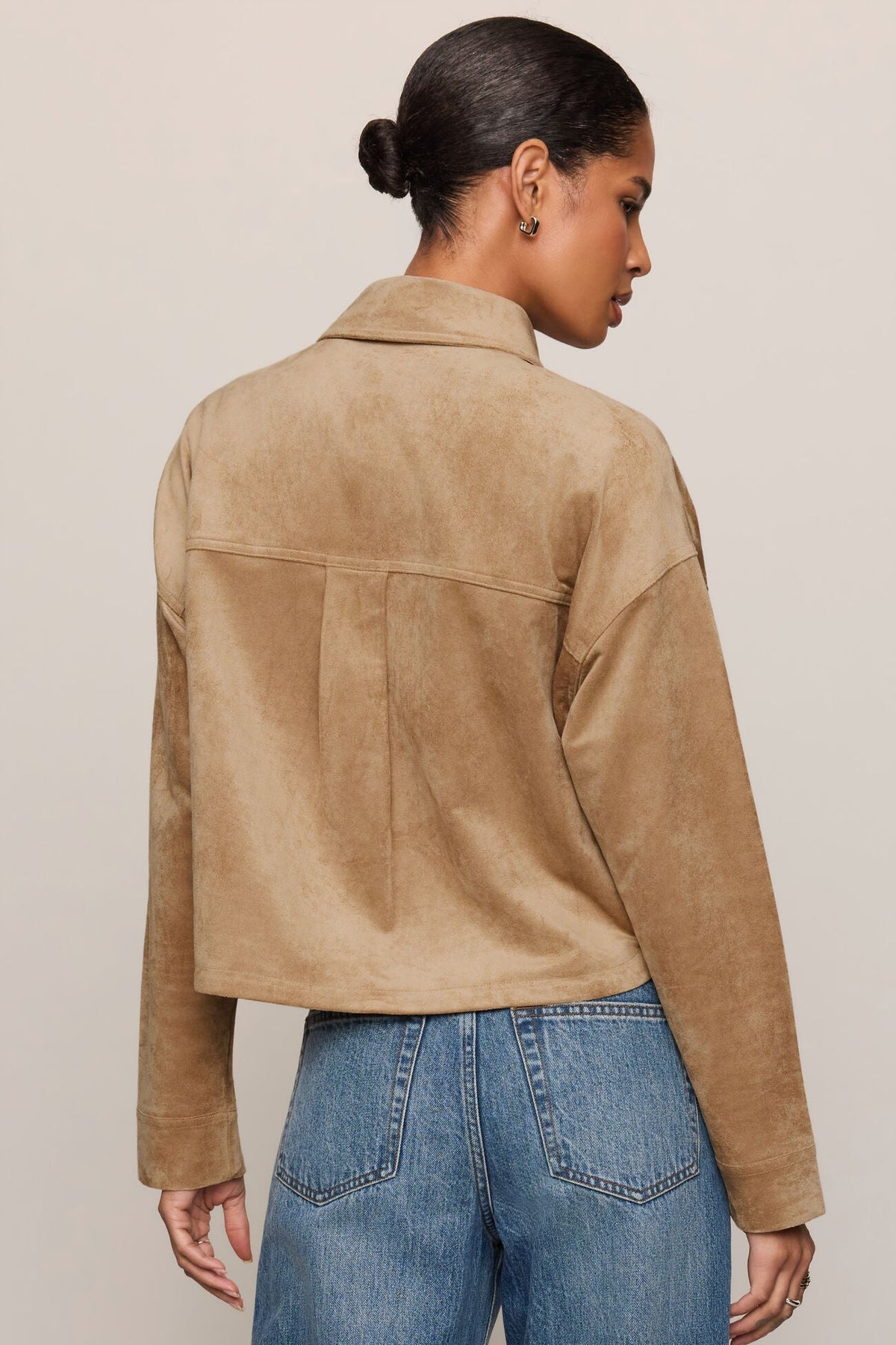 A woman shown from the back wearing a cropped light sand suede jacket and blue jeans against a plain light beige studio background.