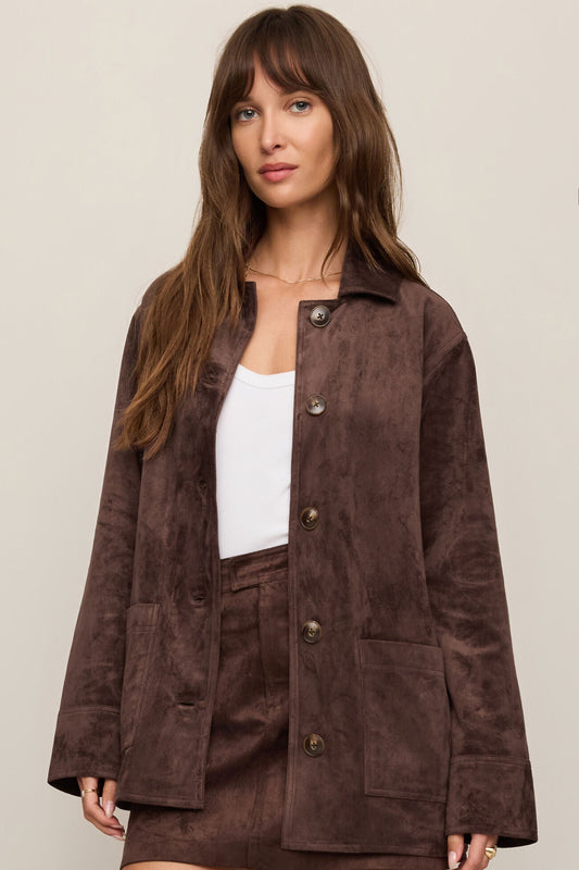 Woman wearing a dark chocolate brown suede jacket over a white top and matching brown shorts on a beige background