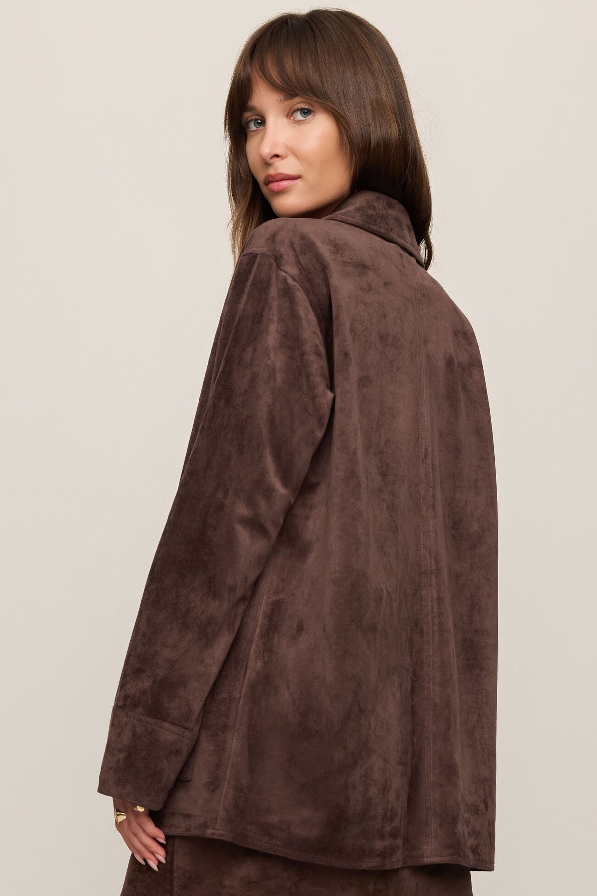 Woman shown from the back wearing a dark chocolate brown suede jacket on a beige background