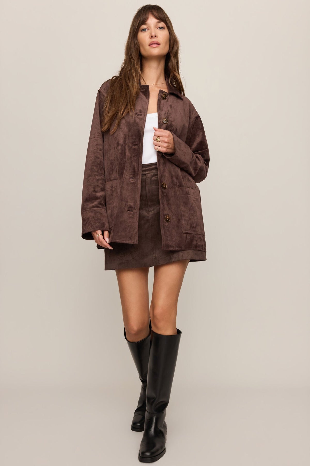 Woman wearing a dark chocolate brown textured jacket and Perri suede mini skirt in dark chocolate with black knee-high boots on a plain background