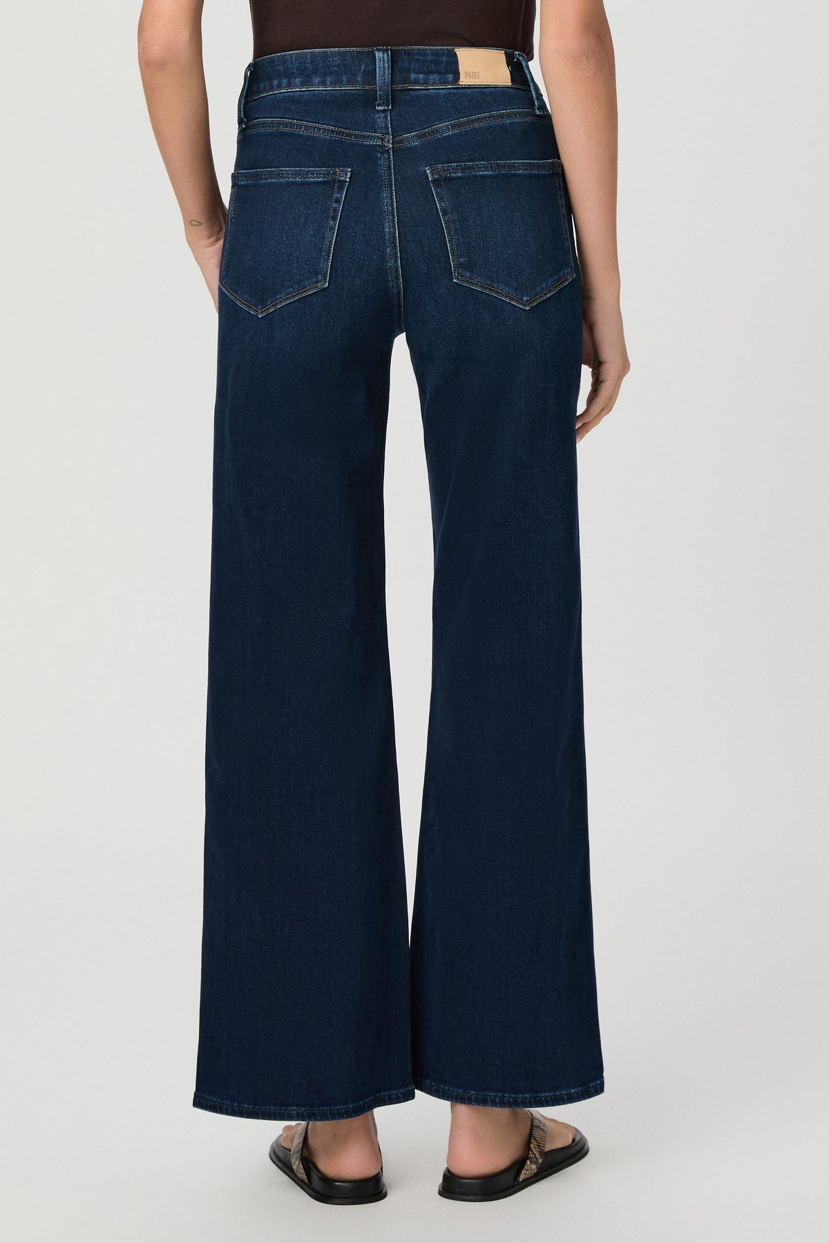 Rear view of a model wearing dark blue amagansett colored wide-leg anessa denim jeans with a straight-to-flared leg and back pocket details, paired with black sandals against a light gray studio background.