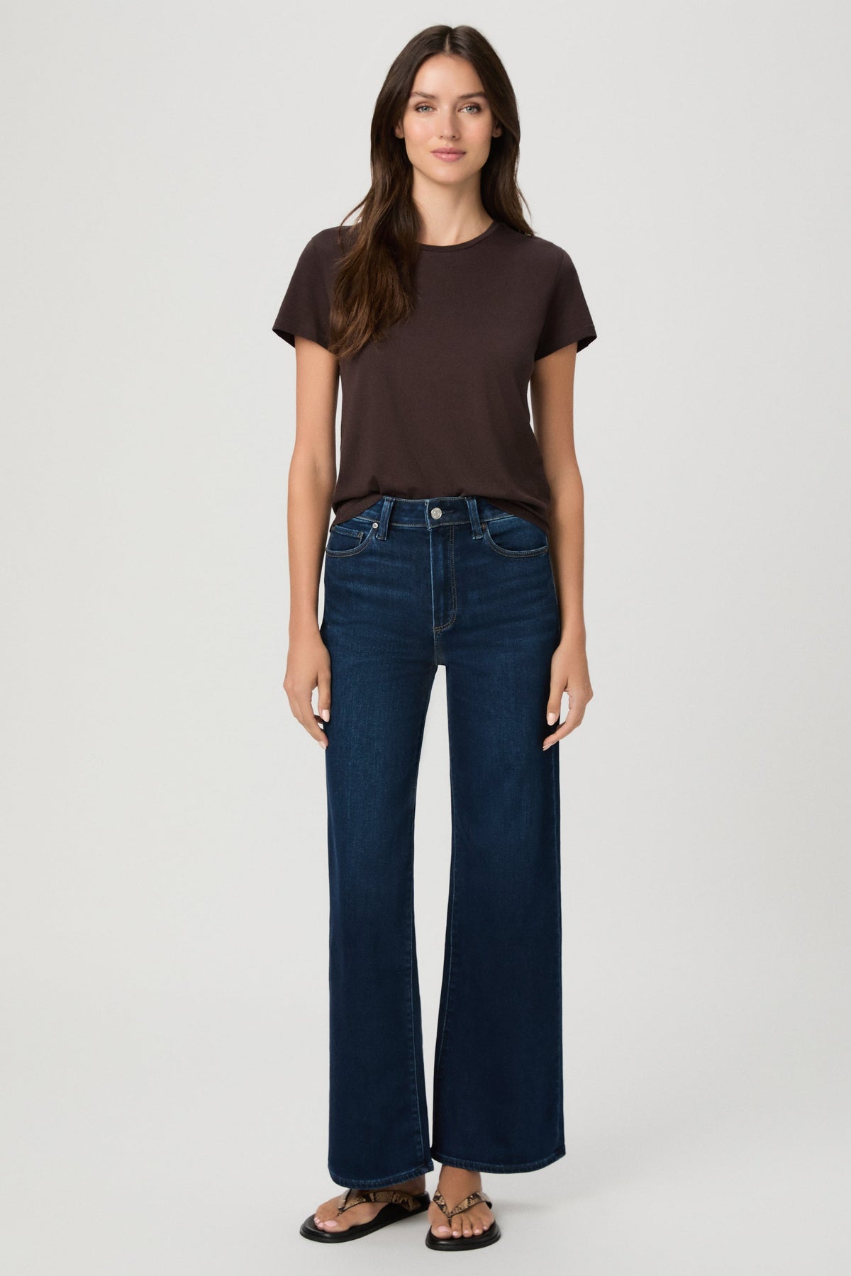 A woman modeling a dark brown short-sleeve t-shirt and dark blue wide-leg jeans in amagansett color against a light gray studio background.