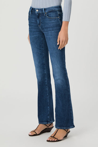 A model wearing a pair of beachline medium-wash blue flare jeans, styled with a light blue ribbed top and black wedge sandals, photographed against a neutral light gray studio background.