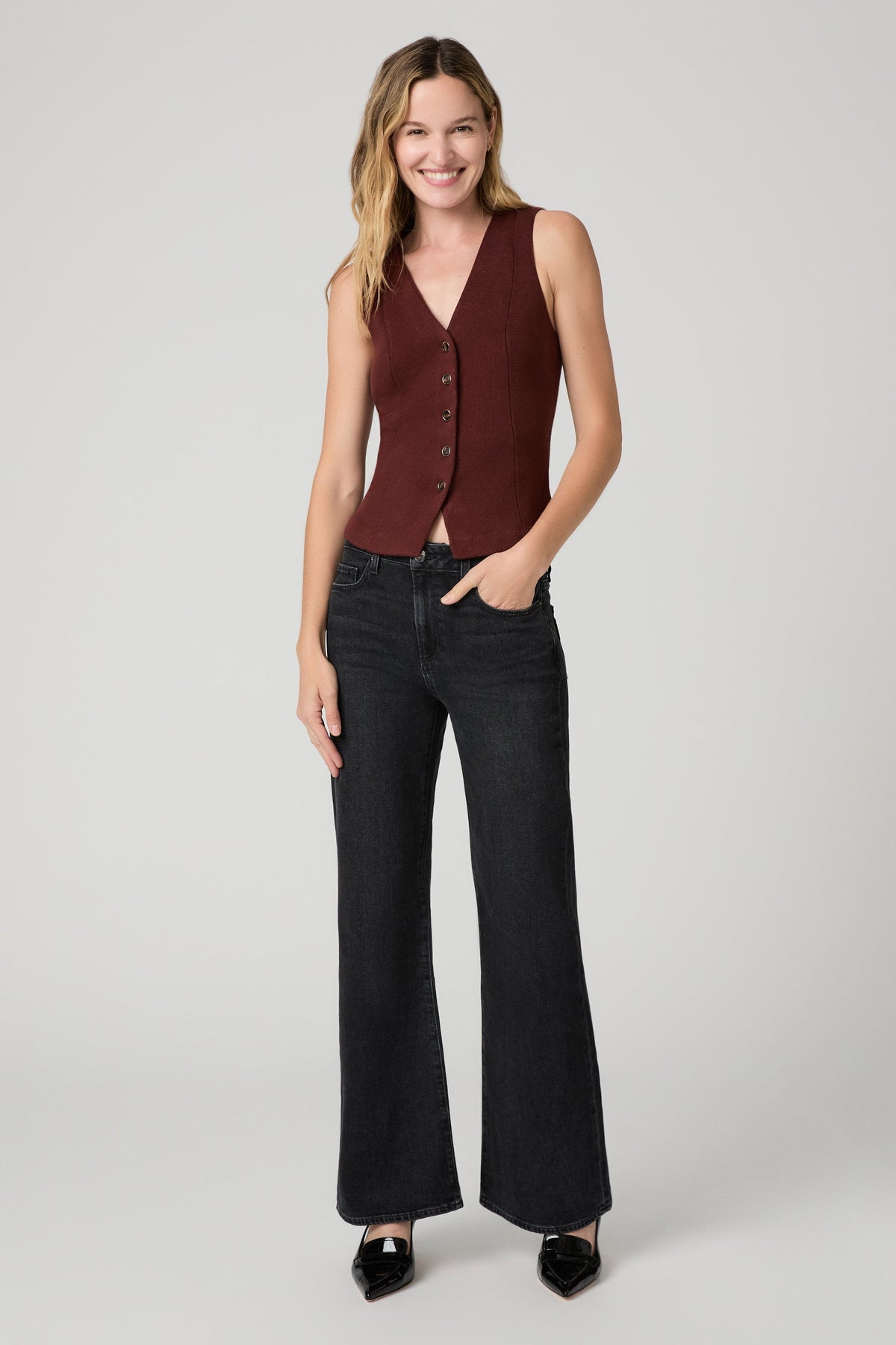 A woman modeling a sleeveless burgundy vest with front buttons over a white top, paired with solstice black wide-leg sofia jeans and black pointed shoes, against a light gray studio background.