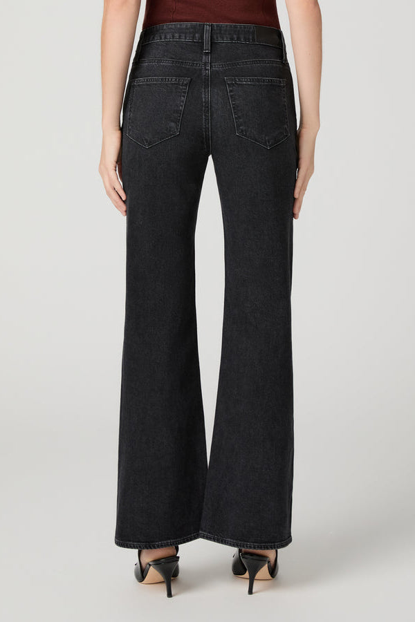 Rear view of a model wearing solstice black wide-leg Sofia denim jeans with a dark wash, paired with black pointed-toe heels against a light gray studio background.