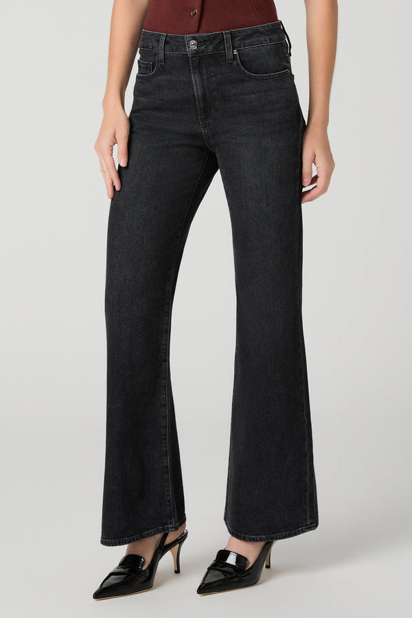A model wearing a pair of solstice black wide-leg flare jeans with a high waist, paired with black pointed-toe mule heels and a dark red top, photographed against a light gray studio background.