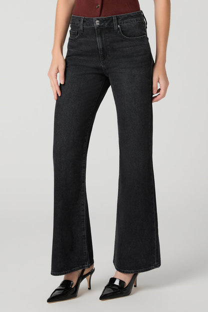 A model wearing a pair of solstice black wide-leg flare jeans with a high waist, paired with black pointed-toe mule heels and a dark red top, photographed against a light gray studio background.