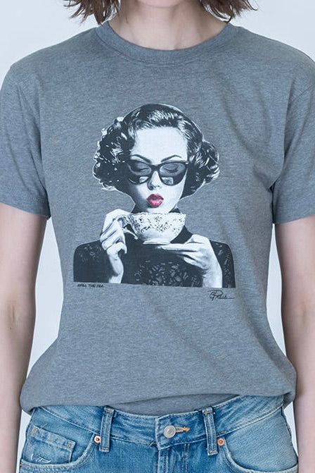 A person wearing a heather gray short-sleeve graphic T-shirt with a black-and-white portrait print of a woman in sunglasses holding a cup of tea, paired with blue jeans against a light gray background.