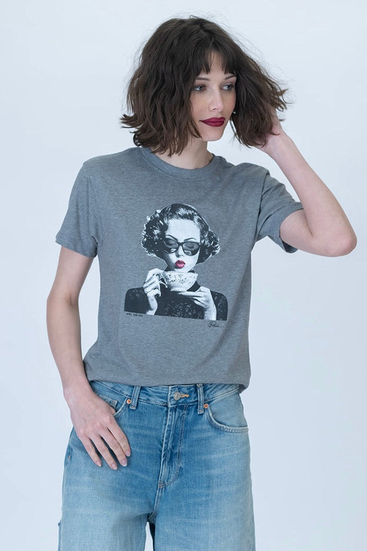 A woman wearing a heather gray graphic t-shirt with a black and white print of a woman drinking tea, paired with light blue jeans, against a light gray studio background.