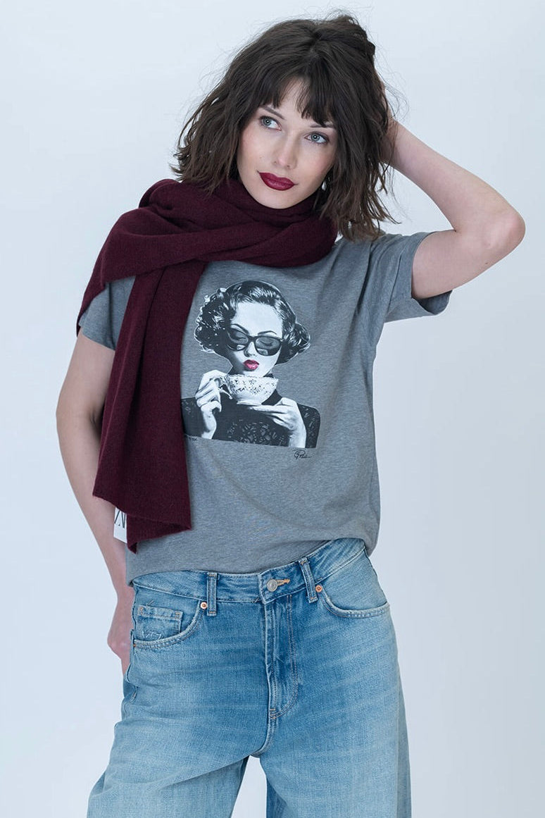 A woman modeling a heather gray spill the tea graphic t-shirt with a black and white portrait print, styled with a dark burgundy scarf and light wash blue jeans against a light gray studio background.