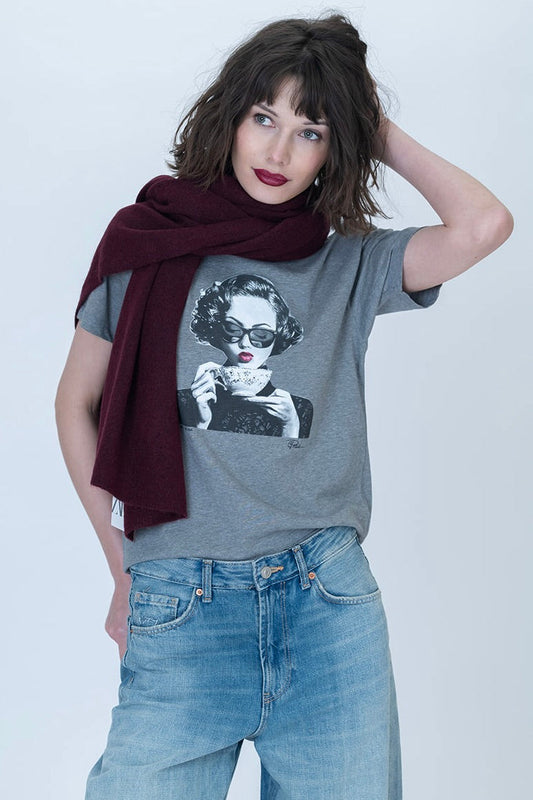 A woman modeling a heather gray spill the tea graphic t-shirt with a black and white portrait print, styled with a dark burgundy scarf and light wash blue jeans against a light gray studio background.