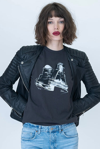 A model wearing a black leather jacket over a black graphic t-shirt with a white high top sneaker illustration, paired with light blue jeans, against a light gray studio background.