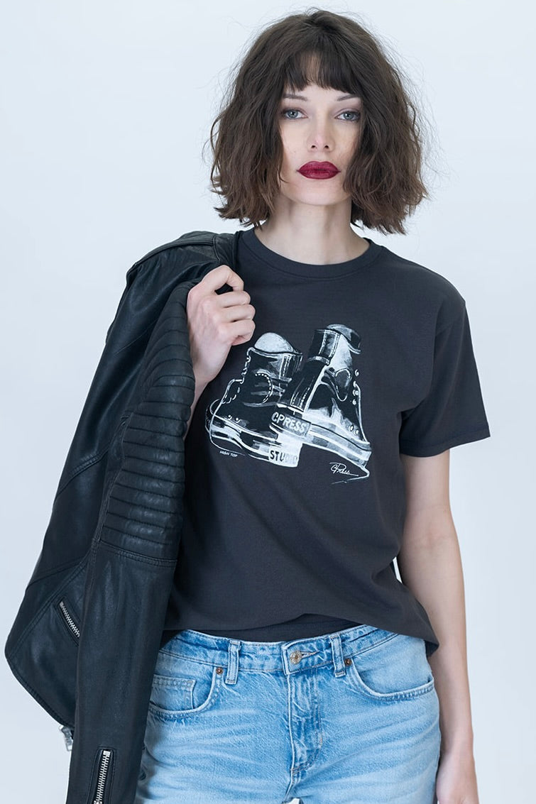 A woman wearing a black graphic t-shirt with a white high top sneaker illustration, styled with light blue jeans and a black leather jacket draped over her shoulder, against a light gray studio background.