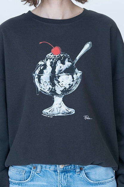 A person wearing a worn black oversized crewneck sweatshirt with a graphic of a vanilla ice cream sundae on the front with a cherry on top, paired with light blue jeans.