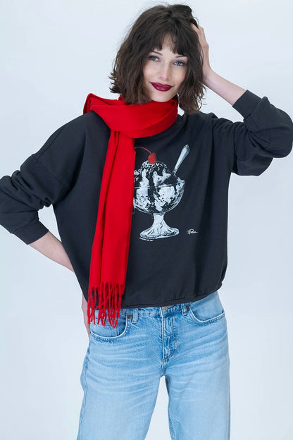 A woman modeling a black sweatshirt with a white ice cream sundae graphic with a cherry on top, styled with a bright red scarf and light blue jeans against a light gray studio background.