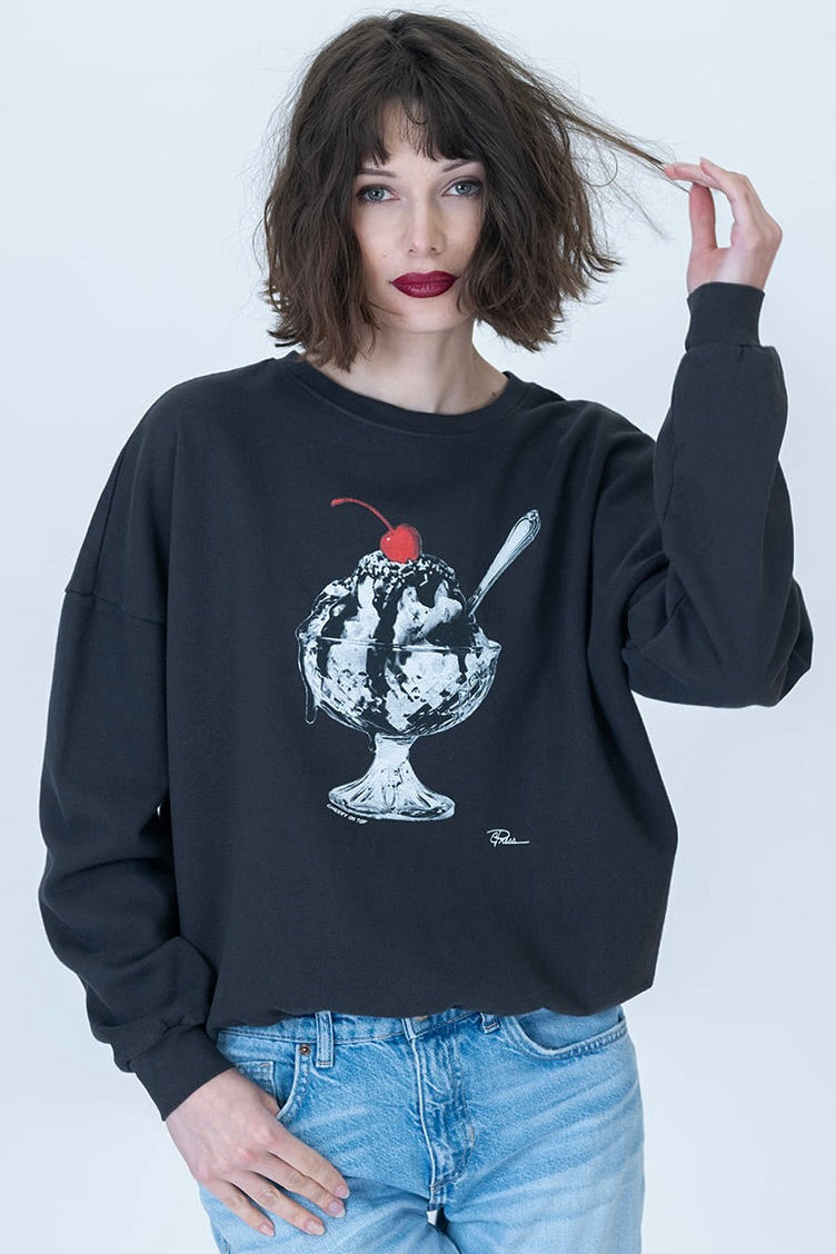 A woman with short dark hair and red lipstick wears a black crewneck sweatshirt featuring a white and gray ice cream sundae graphic with a red cherry on top, paired with light blue jeans, against a plain light gray background.