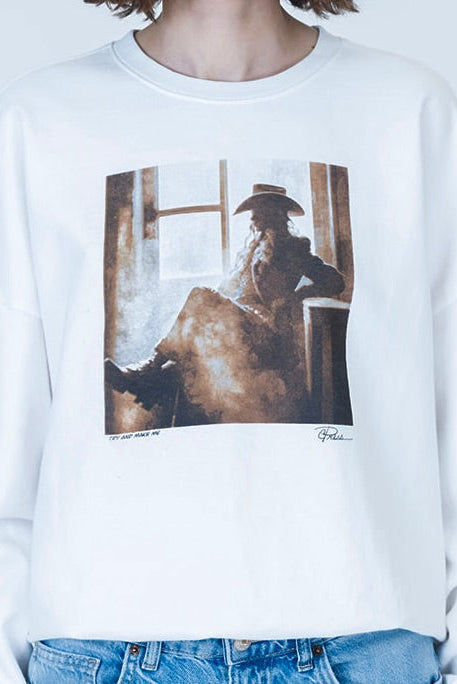 A person wearing a white crewneck sweatshirt with a large square "Try & Make Me Cozy" graphic print on the front showing a cowgirl in a hat, paired with blue jeans against a light gray background.