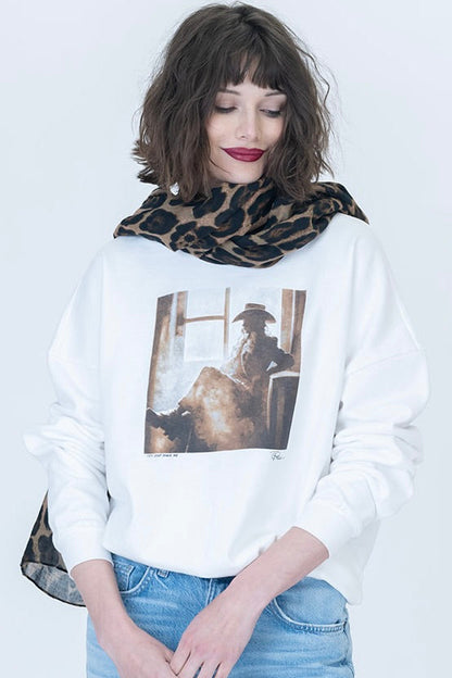 A woman wearing a white sweatshirt with a "Try & Make Me Cozy" large vintage-style western portrait graphic on the front, styled with a leopard-print scarf and blue jeans against a plain white background.
