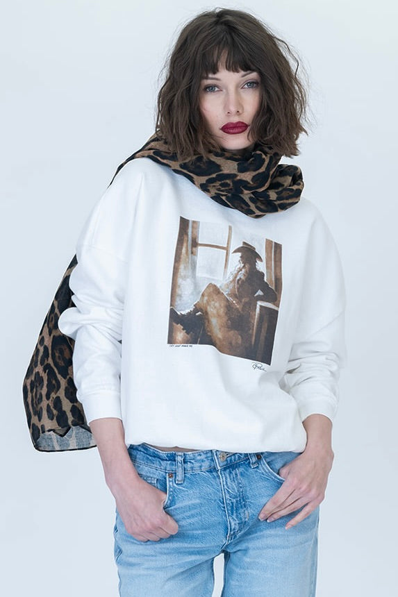 A woman modeling a white crewneck sweatshirt with a vintage-style "Try & Make Me Cozy" graphic print on the front, paired with light blue jeans and a leopard-print scarf, against a plain white background.