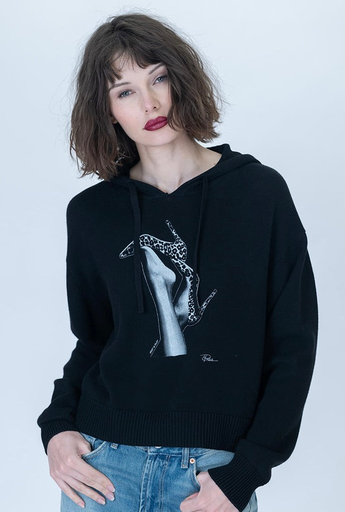 A woman with short brown hair and red lipstick wearing a black pullover hoodie with a large white and black graphic print of leopard heels on the front, paired with blue jeans, against a light gray studio background.