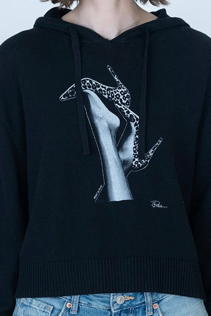 A person wearing a black cropped hoodie with a large graphic print on the front showing legs with high-heeled shoes, with the shoes decorated in a leopard print. The person is also wearing blue jeans against a plain light gray background.