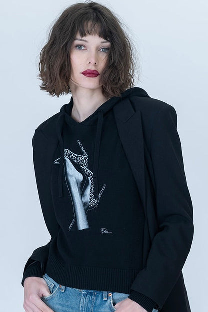 A model wearing a black knit hoodie with a white graphic print of leopard heels on a pair of legs, styled with a black blazer and light-wash distressed jeans against a light gray background.
