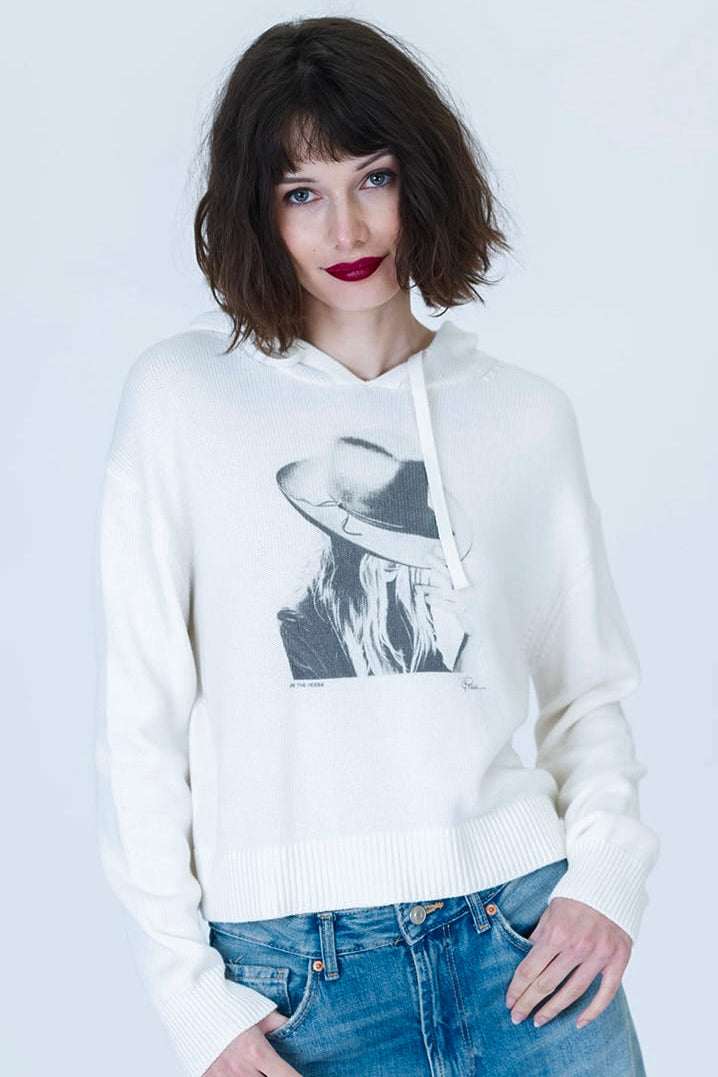 A woman with short dark hair and red lipstick wearing an ivory knit hoodie with a black-and-white "On the Verge Randi" graphic print of a woman in a hat, paired with blue jeans, against a light gray studio background.